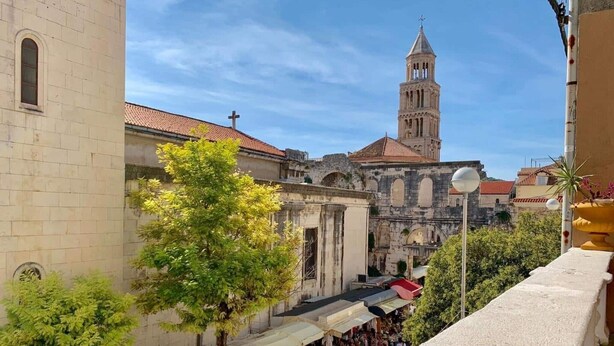 Un edificio histórico con una torre de campanas, un área de mercado con puestos y un árbol en primer plano.