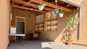 Un patio techado con suelo de baldosas, una mesa de madera, sillas, macetas colgantes y una planta en maceta.