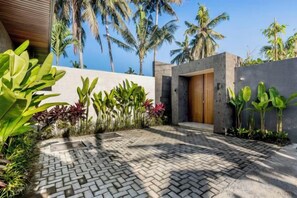 A paved courtyard with tropical plants, a modern building entrance, and palm trees in the background. - Villa Luna by BaliSuperHost (Bali)