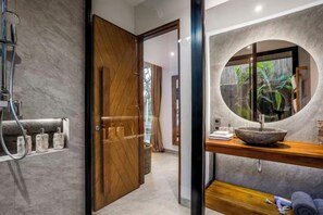 A bathroom with a wooden door, a round mirror, a sink with a bowl, and a towel rack. - Villa Luna by BaliSuperHost (Bali)