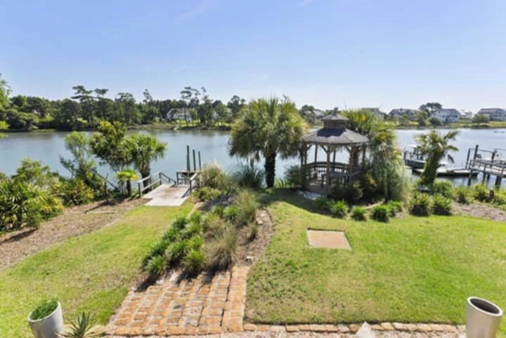 Um gazebo à beira de um lago, com gramado, um caminho de tijolos e um pequeno deck.