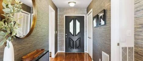 A hallway with a wooden floor, a mirror, a door with a decorative design, and a wall-mounted picture frame.
