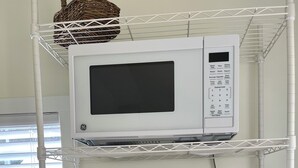 Un coin cuisine avec un four à micro-ondes GE posé sur une étagère blanche, une machine à café Keurig, des récipients en acier inoxydable, une assiette avec des tomates et des raisins, et un vase avec des tournesols.