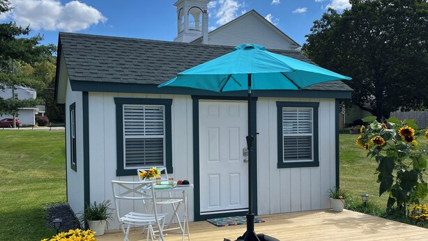 Un petit bâtiment d’un seul étage, à l’extérieur blanc et aux bordures sombres, avec un patio couvert agrémenté d’un parasol turquoise, et une table et une chaise blanches disposées à l’extérieur.
