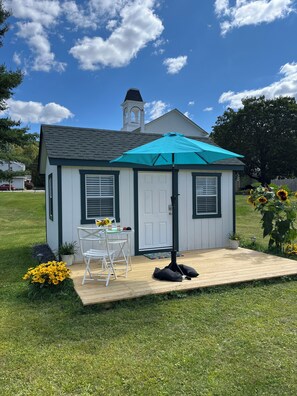 A small, single-story building with a white exterior and dark trim, a covered patio with a turquoise umbrella, and a white chair and table set up outside. - Oneonta/Cooperstown Baseball CharmingCottage at the church in Mount Vision. (Mount Vision)