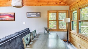 Ein Holzblockhaus-Interieur mit einem Esstisch, Bänken, einem Schild, das zum See weist, und einem Fenster mit Blick auf Bäume.