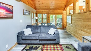 Ein gemütliches Wohnzimmer mit grauem Sofa, Holzwänden und großen Fenstern.