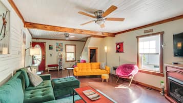Un salon chaleureux avec un canapé vert, un divan orange vif et un fauteuil rose. On peut voir un ventilateur de plafond et une cheminée.