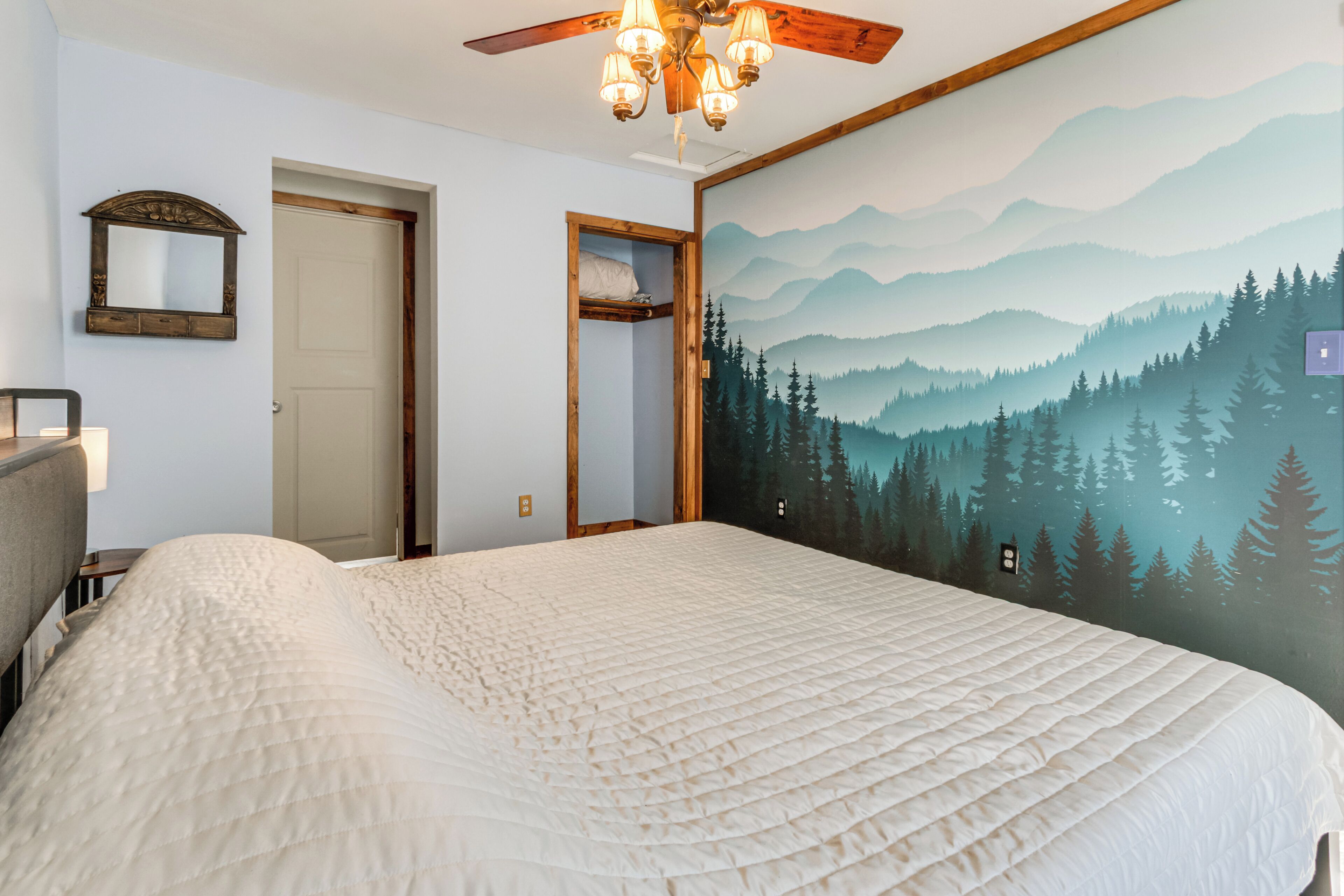 Une chambre avec un grand lit, un ventilateur de plafond, un miroir fixé au mur et une fresque représentant une forêt.
