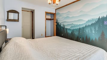 Une chambre avec un grand lit, un ventilateur de plafond, un miroir fixé au mur et une fresque représentant une forêt.