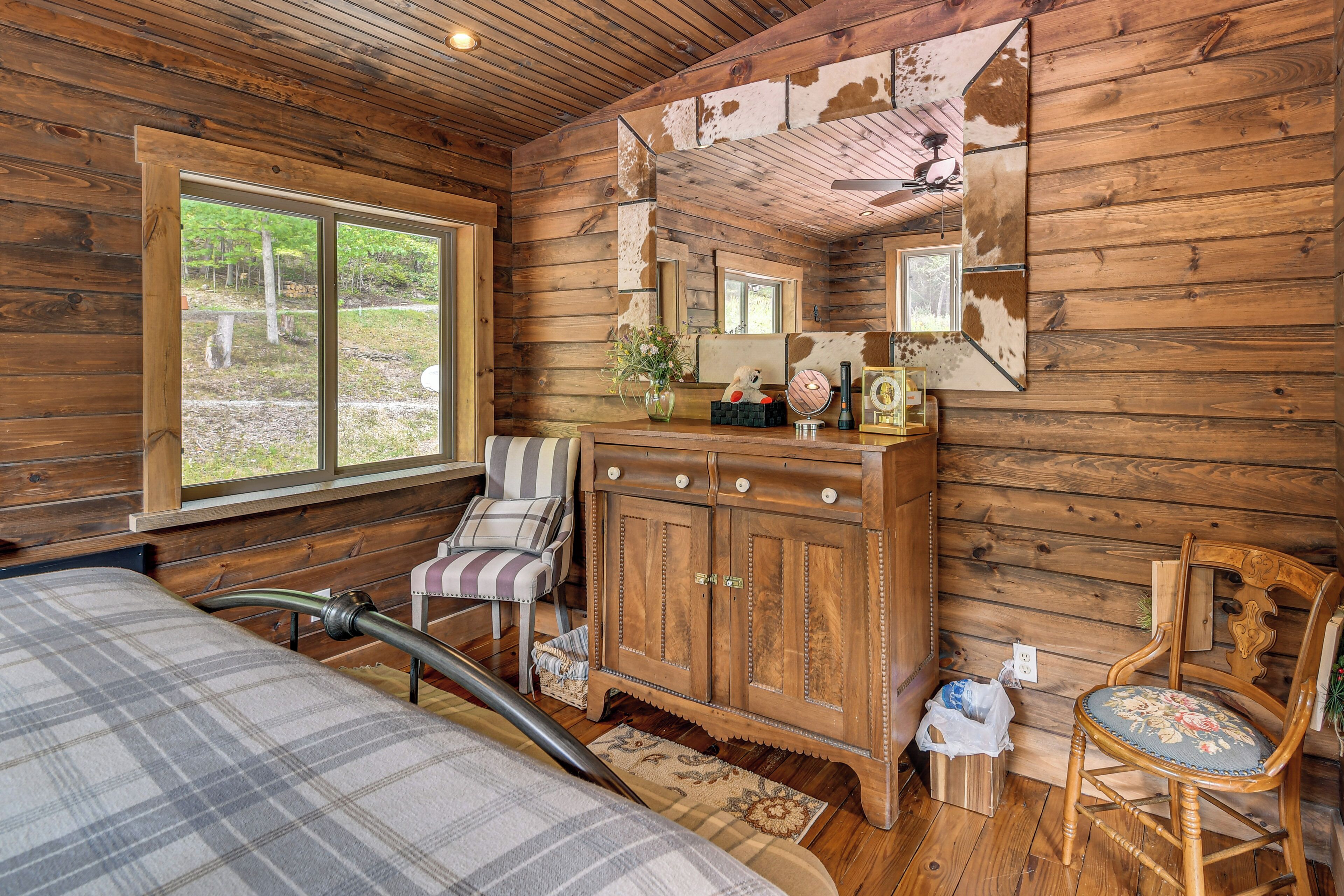A wooden cabin interior with a large window, a plaid bed, a wooden cabinet, and a chair.