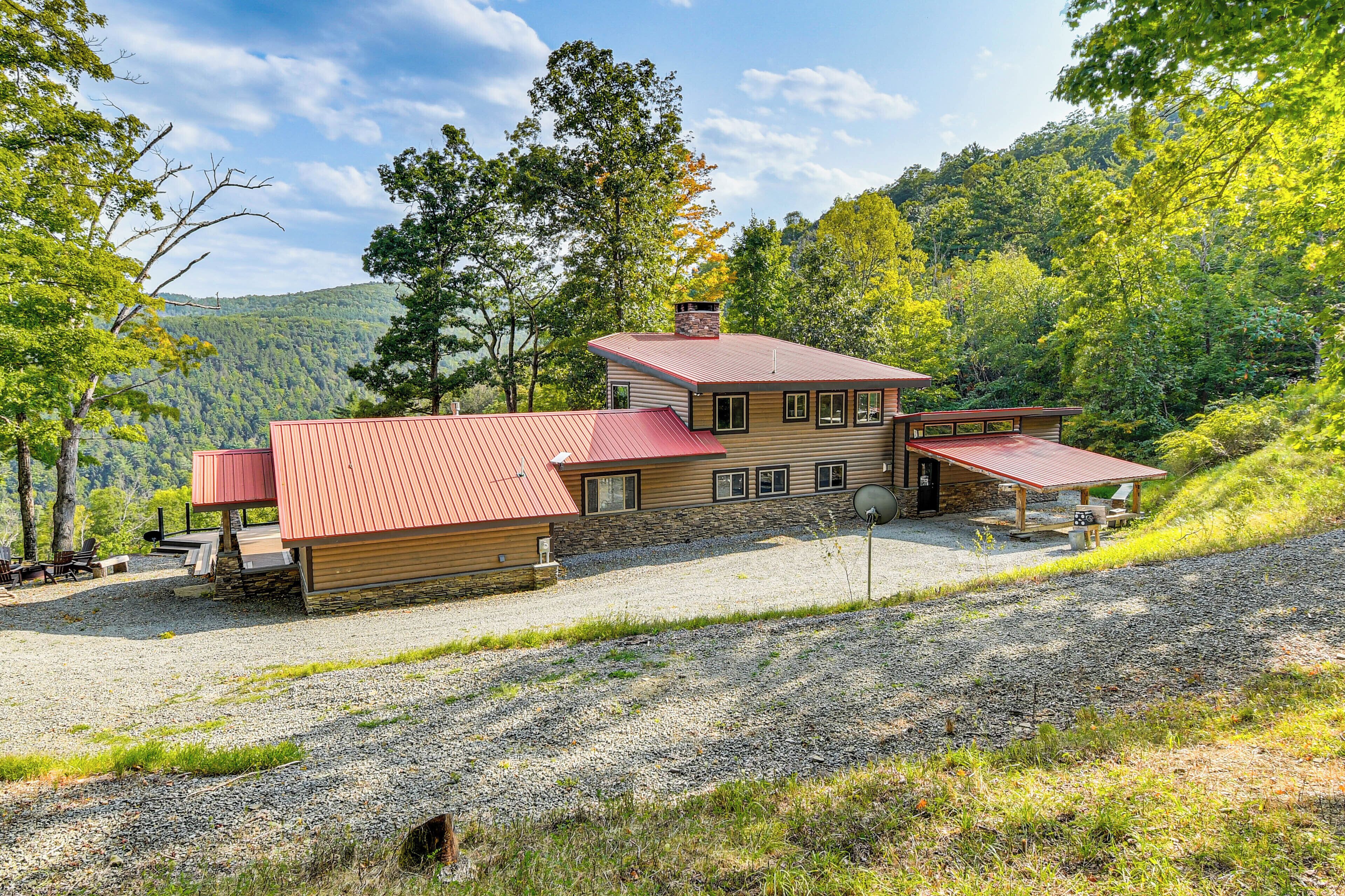 A two-story cabin with a red roof, a stone chimney, and a gravel driveway.