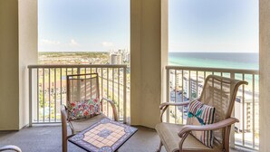 A balcony with two wicker chairs, one with a floral cushion and the other with a striped pillow, overlooking a cityscape and ocean.