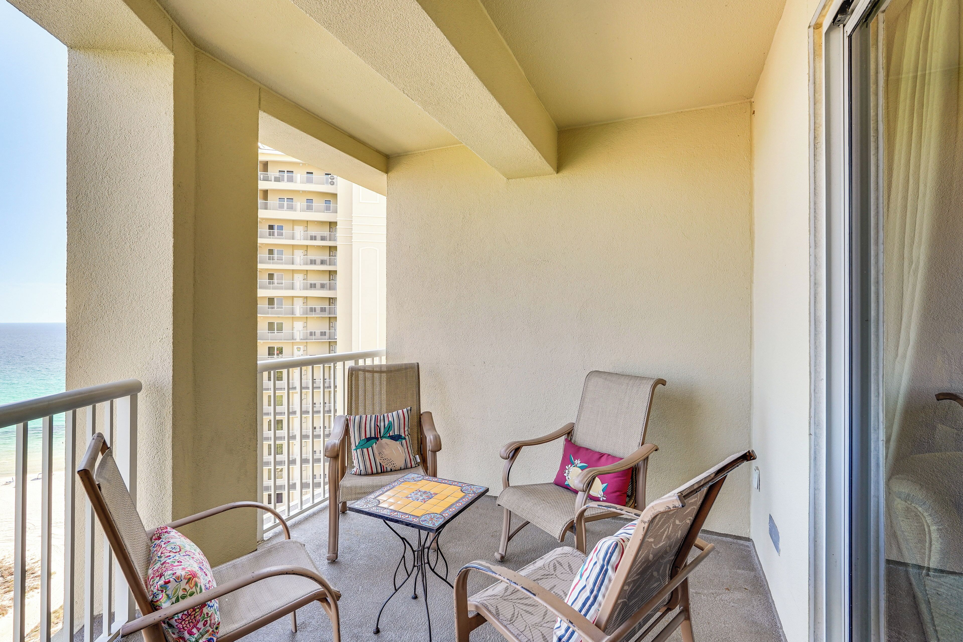 Ein Balkon mit Korbstühlen und einem kleinen Tisch, mit Blick auf einen Strand und ein Hochhaus.