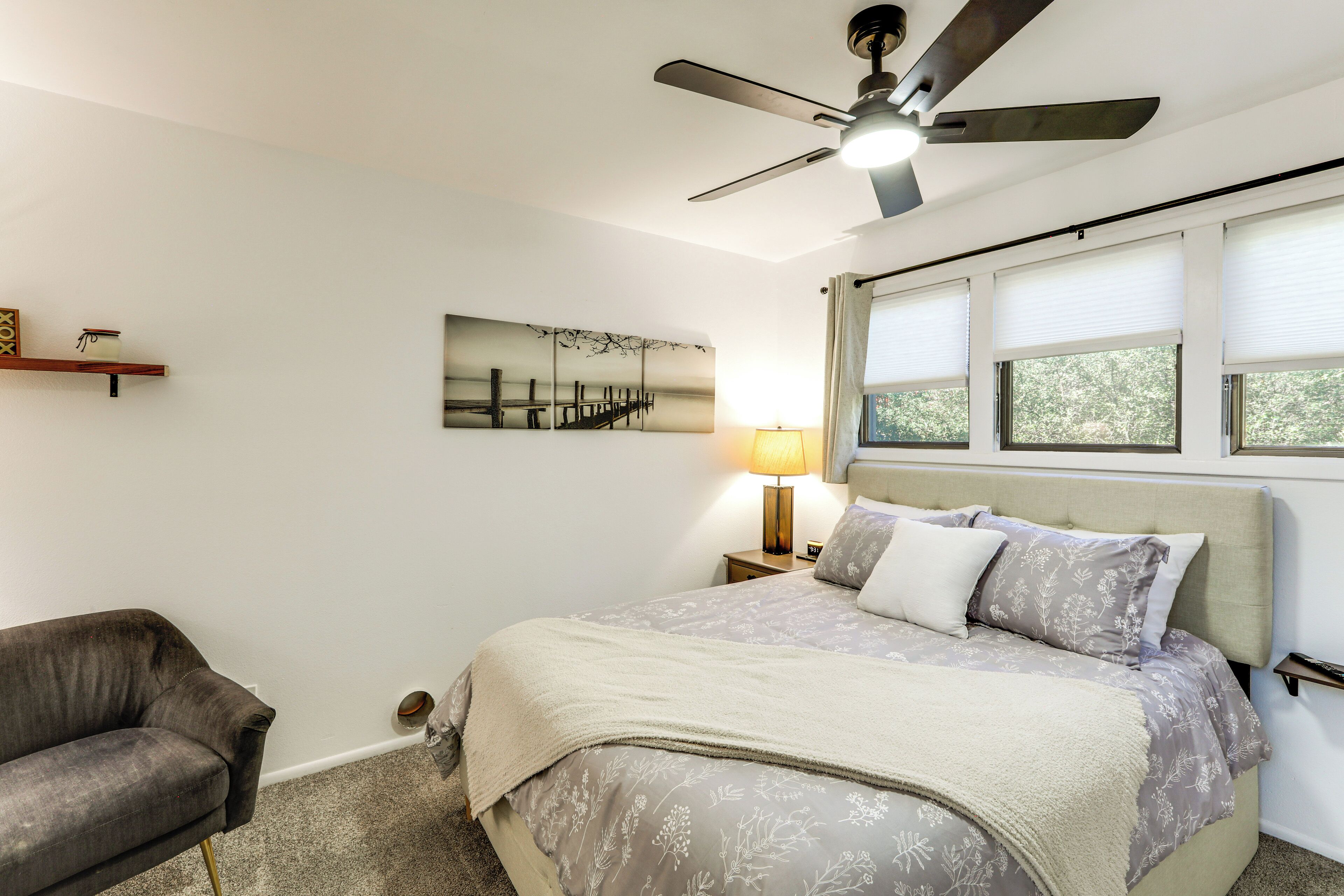 A bedroom with a bed, a ceiling fan, a nightstand, a lamp, a chair, and a window with blinds.