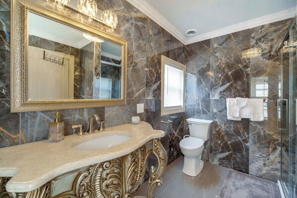Un baño con paredes de mármol, un espejo con marco dorado, un lavabo, un inodoro y una cabina de ducha de vidrio.