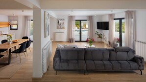 Una sala de estar moderna con un sofá gris, comedor con mesa y sillas de madera, y un televisor grande montado en la pared.
