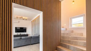 A modern interior with a grey sofa, a flat-screen TV, and a staircase with a window. - Villa Neoma – Tranquil Seaside Escape with Heated Pool and Breathtaking Sea Views (Linardići (Krk))