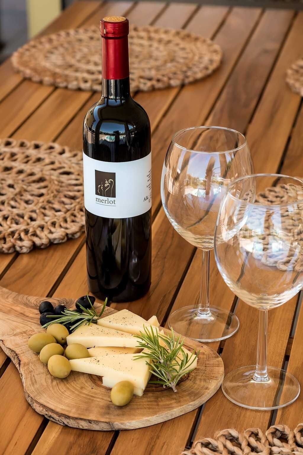 A bottle of Merlot wine, two glasses, a wooden cutting board with cheese and olives, and a woven placemat on a wooden table.