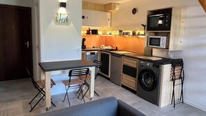 Una cucina compatta con forno, microonde e lavatrice integrati. C'è un'area pranzo con tavolo e sedie, e un divano grigio.