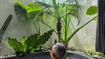 A person seated in a bathtub with large green plants in the background.