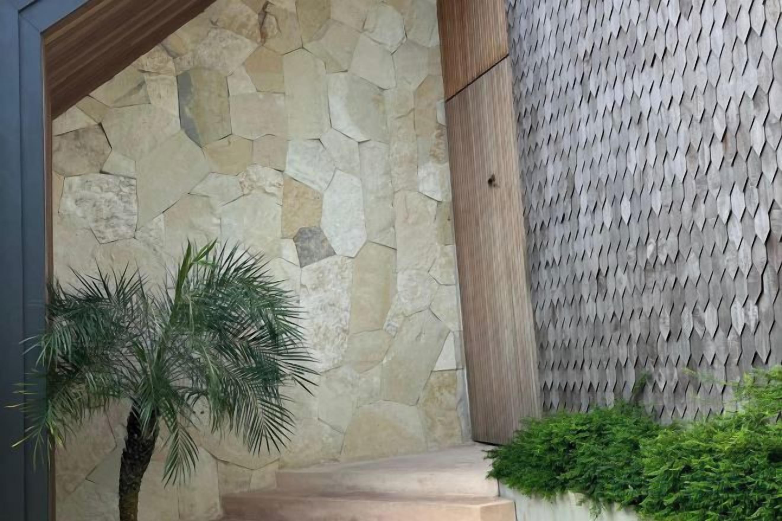 A stone wall with a textured pattern, a palm tree, and a green hedge.
