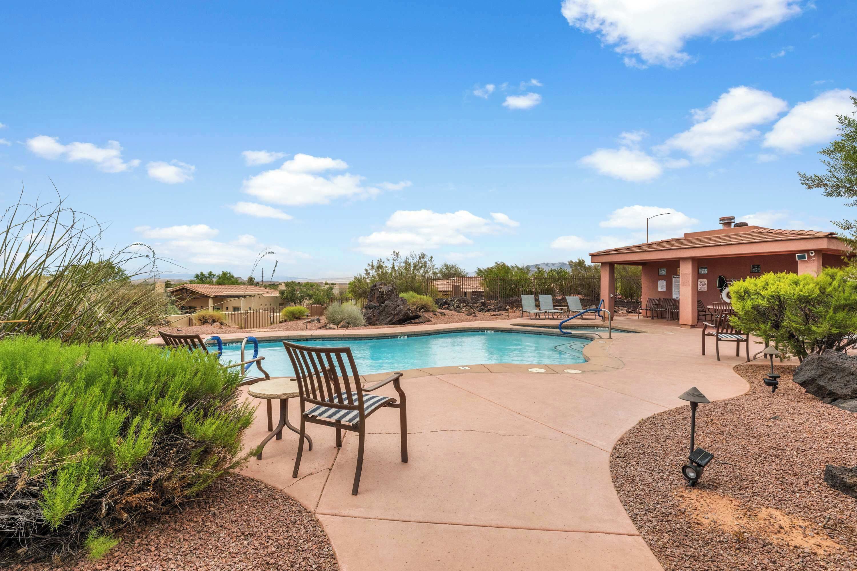 A pool area with lounge chairs and a patio table, surrounded by landscaping and a building with a red roof.