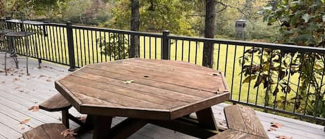 Um deck de madeira com mesa e bancos, rodeado por um guarda-corpo preto, com vista para uma área arborizada.
