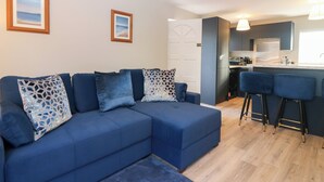 Una sala de estar moderna con un sofá modular, una zona de cocina con taburetes de bar y una puerta que da a otra habitación.