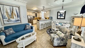 Un grand salon avec un canapé bleu, un fauteuil à motifs et un transat blanc pour bébé. On aperçoit une table à manger avec des chaises à l’arrière-plan.