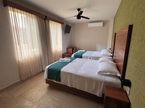 Een hotelkamer met twee bedden, een plafondventilator, een airconditioningapparaat, een aan de muur bevestigde televisie en een groot raam met gordijnen.