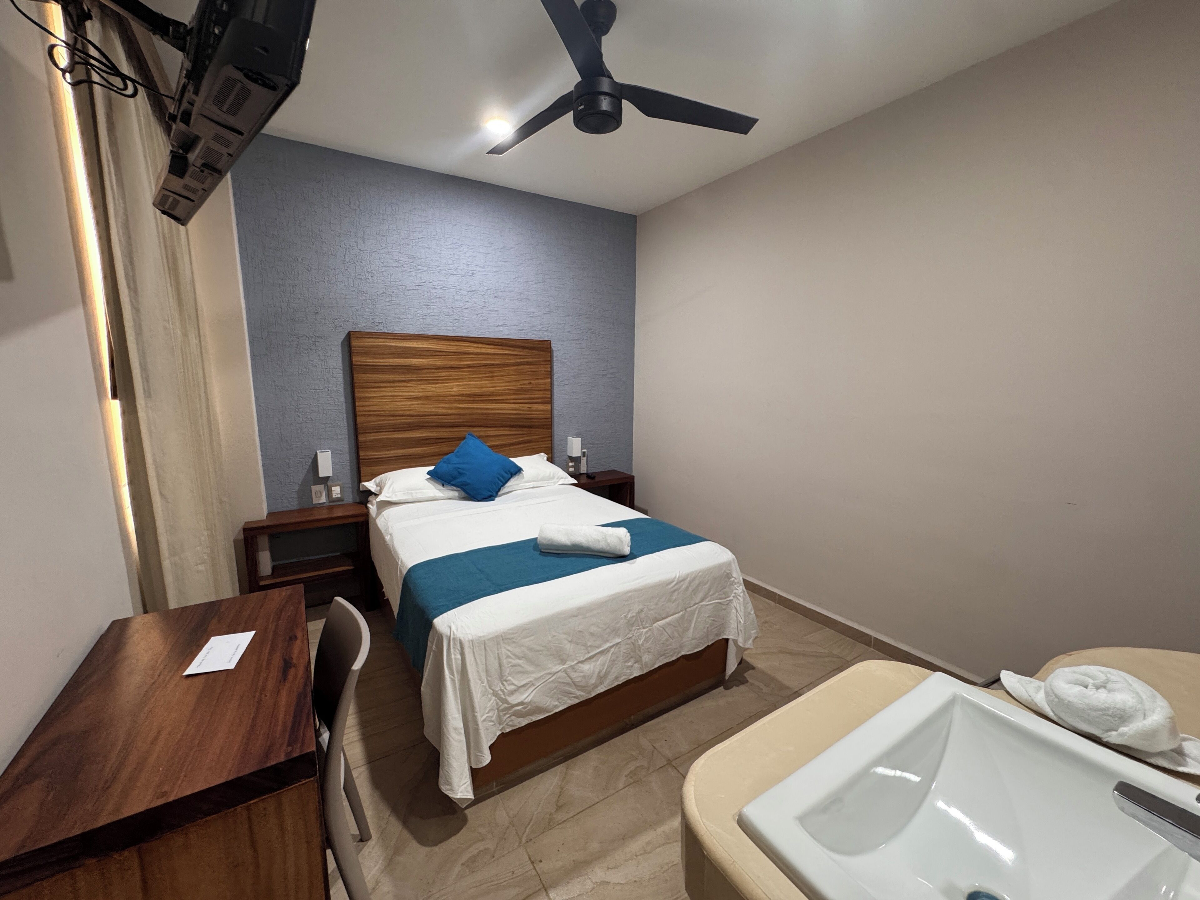 A hotel room with a bed, a desk, a chair, a bathroom with a sink and mirror, and a ceiling fan.