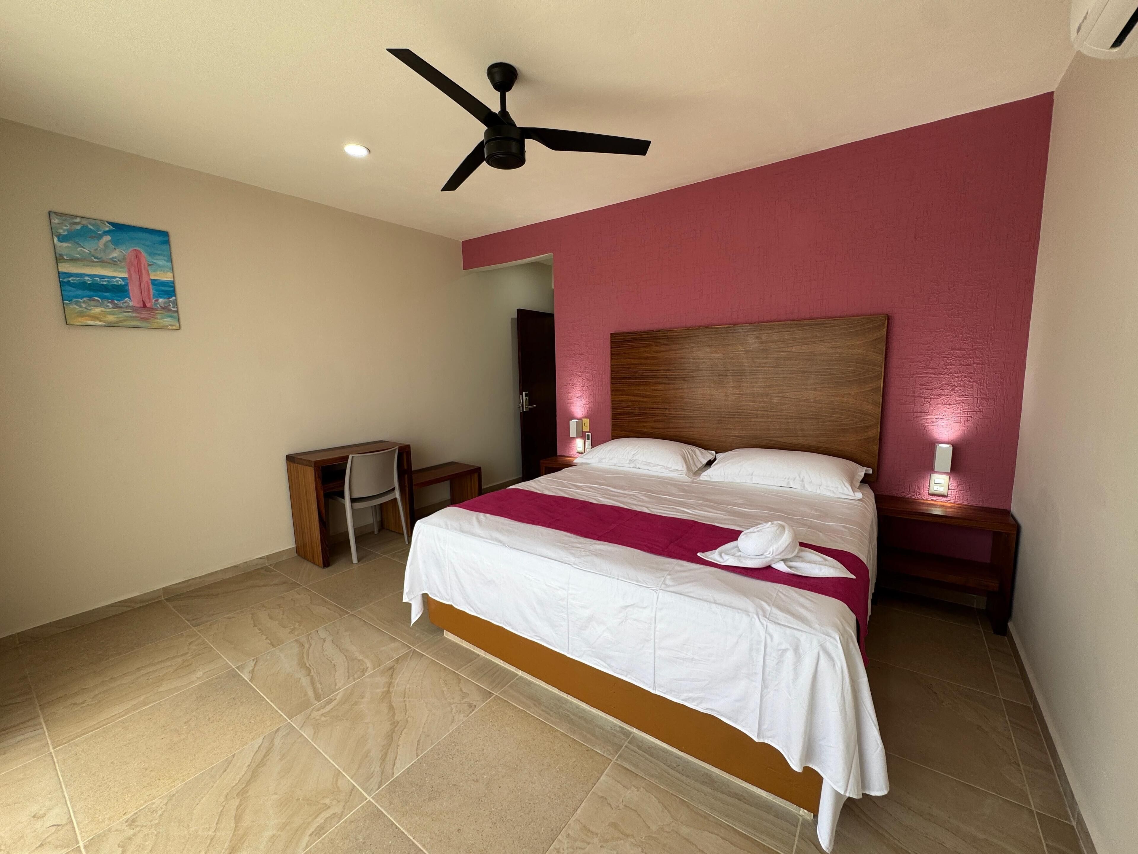 A hotel room with a bed, a desk, a chair, a ceiling fan, and a painting on the wall.