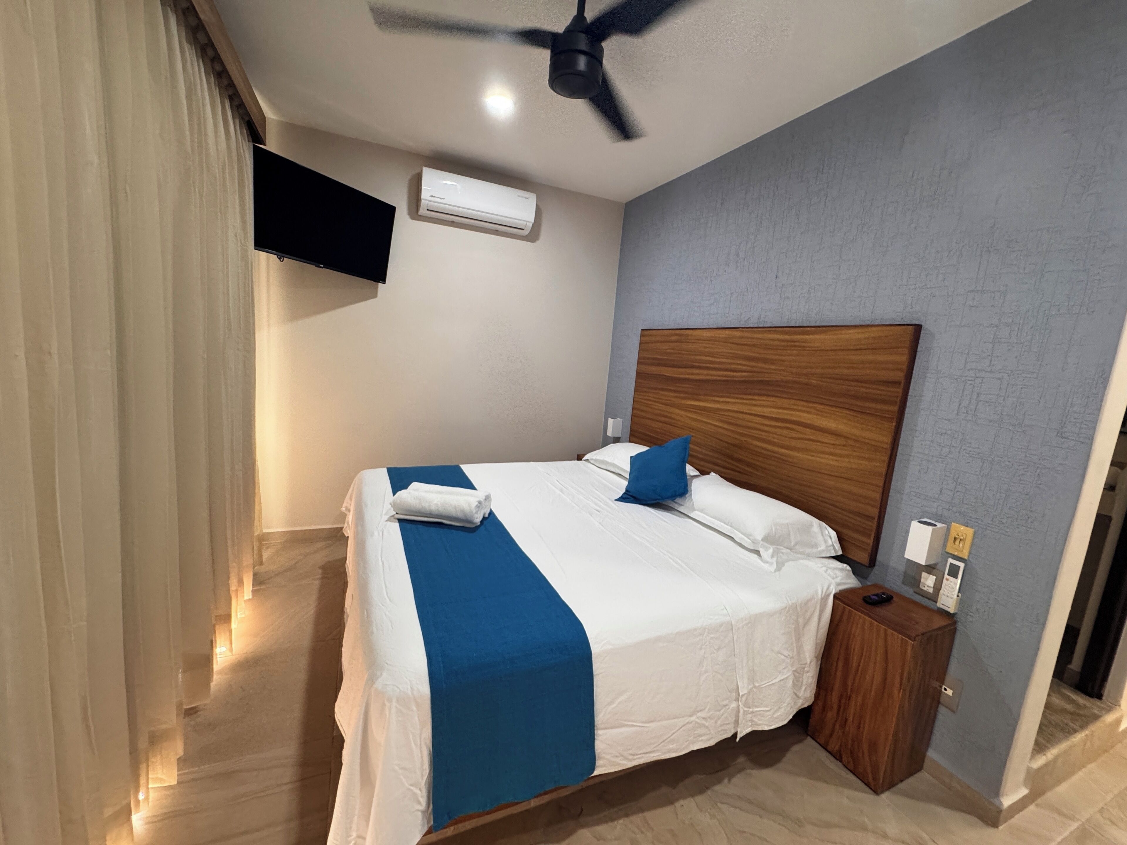 A hotel room with a bed, a wooden headboard, a nightstand, a TV mounted on the wall, and an air conditioning unit.