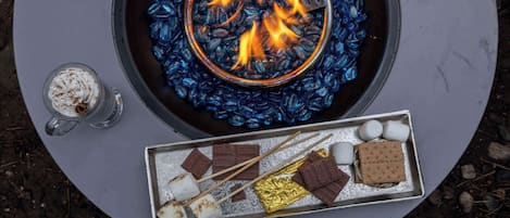 Un feu de camp avec un plateau en métal contenant des guimauves, du chocolat et des biscuits Graham.