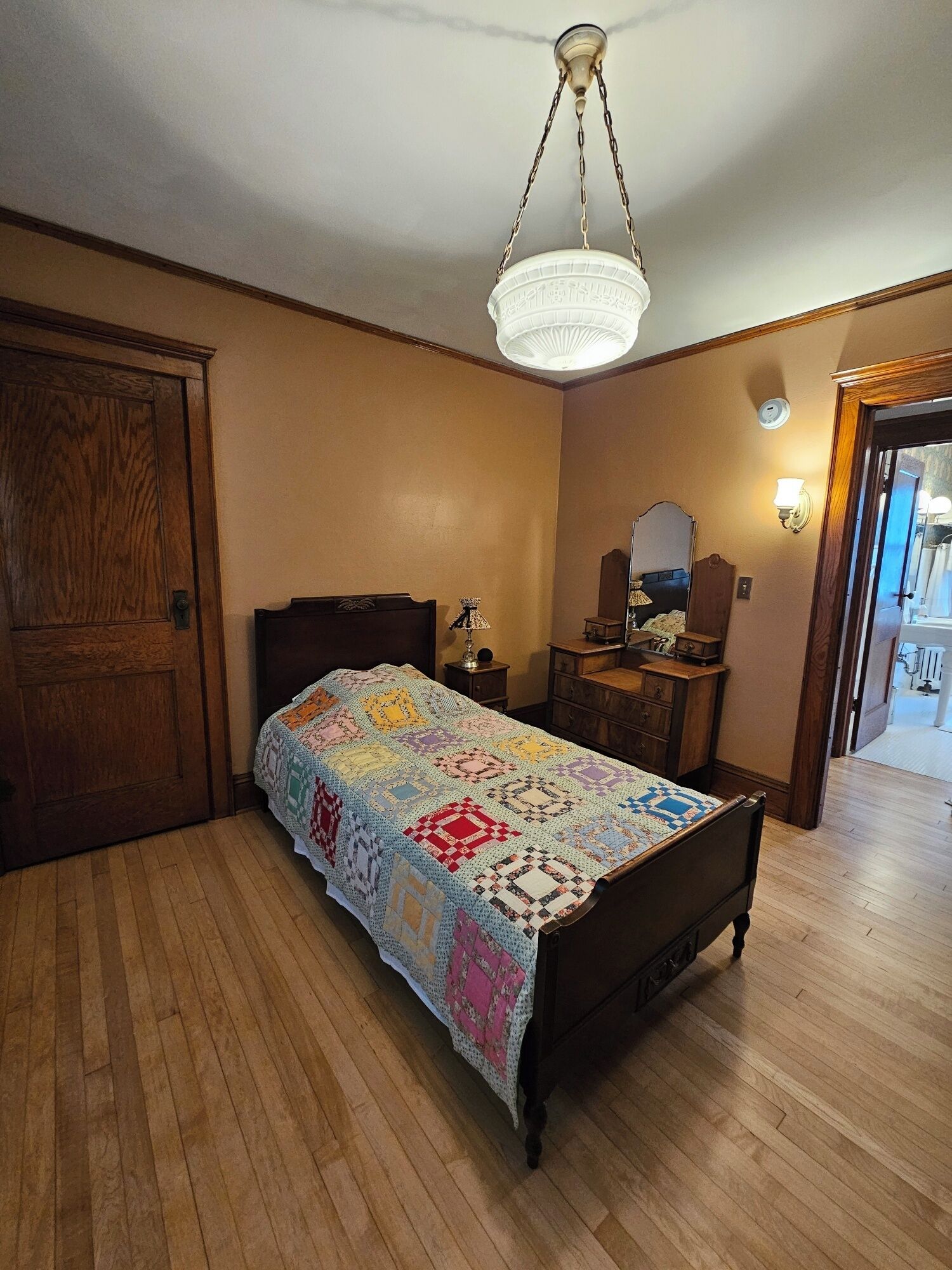 A bedroom with a wooden floor, a bed with a colorful quilt, a wooden dresser, a nightstand, a mirror, and a door.