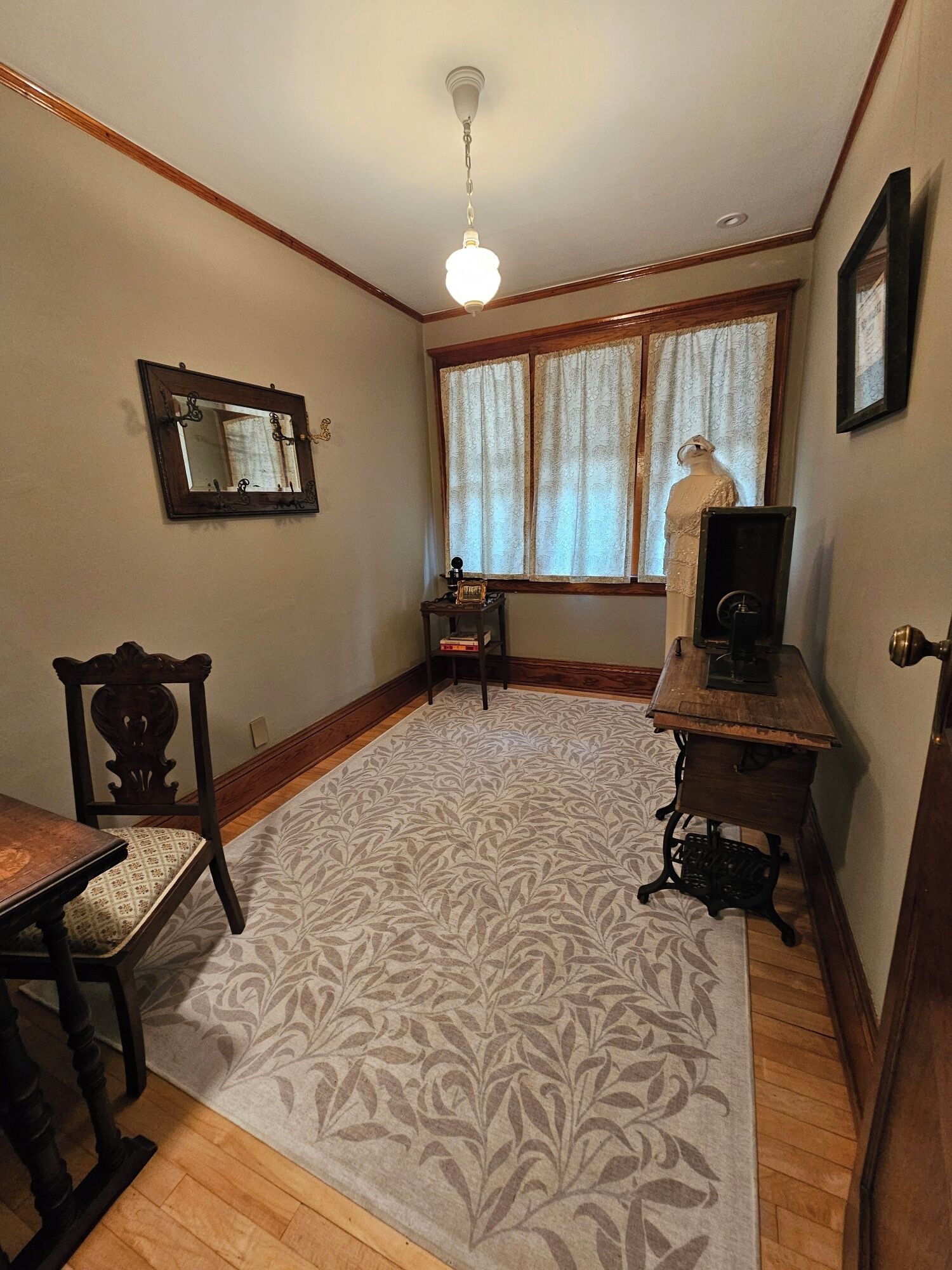 A room with a wooden floor, a patterned rug, a wooden chair, a table, a mirror, a mannequin, a sewing machine, and a window with curtains.