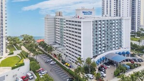 A high-rise building with a sign reading "Marco Polo Beach Resort." It features multiple floors with balconies, a parking area, and a covered entrance with blue canopies.