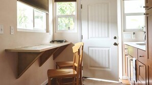 Een keuken met een houten aanrecht, een houten stoel, een witte deur en een raam met jaloezieën.
