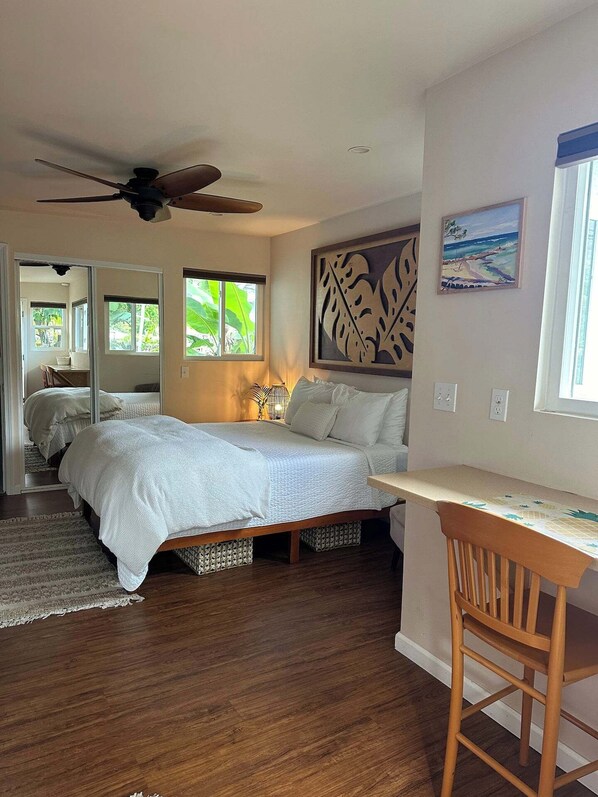 A bedroom with a bed, a desk, and a chair. There is a ceiling fan, a window with a view of greenery, and a painting on the wall.