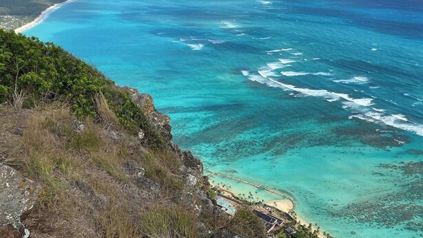 A coastal landscape with clear turquoise waters, a sandy beach, and a rocky cliff with sparse vegetation.