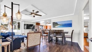A modern living room with a blue sofa, zebra-patterned chairs, a dining area with wooden chairs, a ceiling fan, and a large painting of a beach.