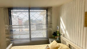 Un dormitorio con una cama, una ventana con persianas, una mesita de noche con una planta y un panel de pared vertical.