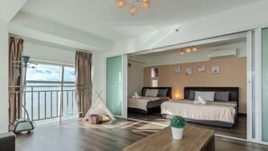 A modern hotel room with a large sliding glass door leading to a balcony, a bed with a white and gray color scheme, a small seating area with a wooden table, and a potted plant. - Maritime Seaview Comfy Suite in Penang with TVbox.Pool.Wifi (George Town)