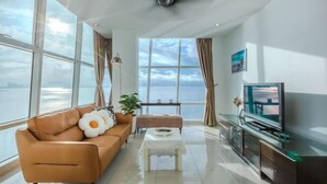 A modern living room with a sofa, a coffee table, a TV, and large windows overlooking a body of water. - Maritime Seaview Comfy Suite in Penang with TVbox.Pool.Wifi (George Town)