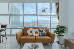 A modern living room with a leather sofa, a coffee table with remotes, a large window with a city view, and a potted plant. - Maritime Seaview Comfy Suite in Penang with TVbox.Pool.Wifi (George Town)