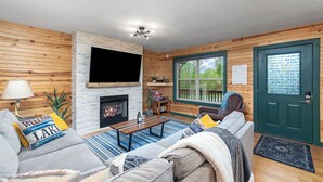 A cozy living room with a fireplace, wooden walls, a television, and a green door. - Wren Cabin - Private Hot Tub, Fireplace, and Lake View (Forsyth)