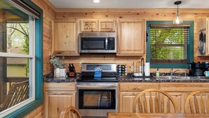 A kitchen with wooden cabinets, a stainless steel microwave, an oven, and a window with blinds. - Wren Cabin - Private Hot Tub, Fireplace, and Lake View (Forsyth)
