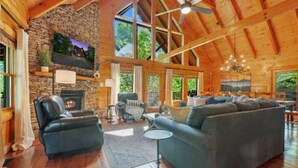 A spacious living area with a stone fireplace, leather chairs, a sofa, and large windows offering natural light and views of the outdoors. - Rustic Cove: Views, Hot Tub, Theater & Game Room! (Sevierville)