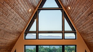 A spacious room with a high ceiling, large windows offering a view of a forested landscape, and a wooden floor.